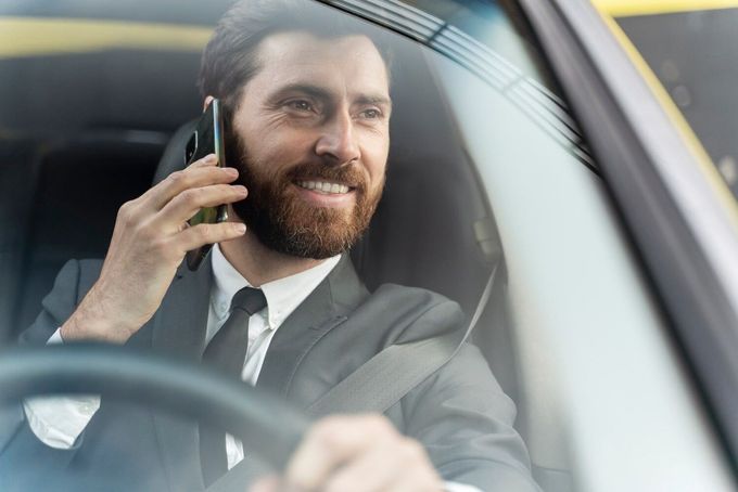 Un hombre sonriente con traje de negocios conduce un coche mientras habla por teléfono inteligente.
