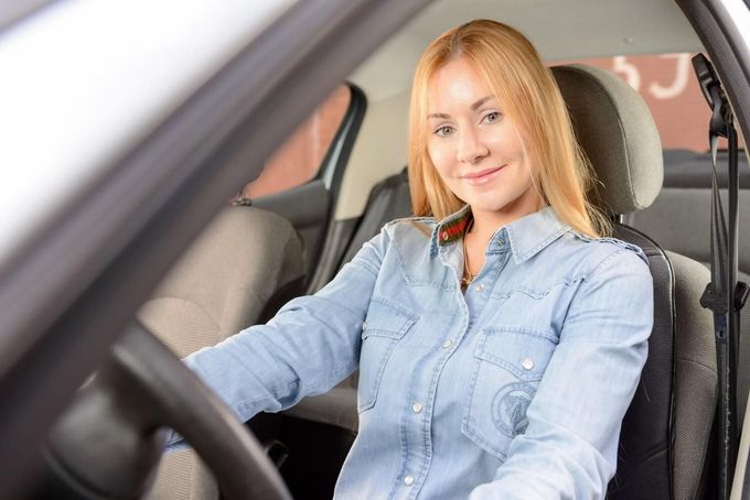 Una persona sonriente, vestida con una camisa vaquera, está sentada en el asiento del conductor de un coche.