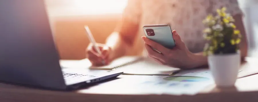 Una persona escribe junto a un portátil mientras sostiene un teléfono inteligente, con una pequeña planta en maceta sobre el escritorio.