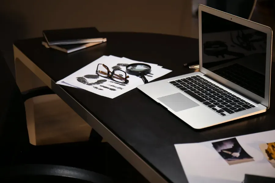 Un portátil y papeles sobre un escritorio oscuro con gafas, un cuaderno y fotos impresas cerca.