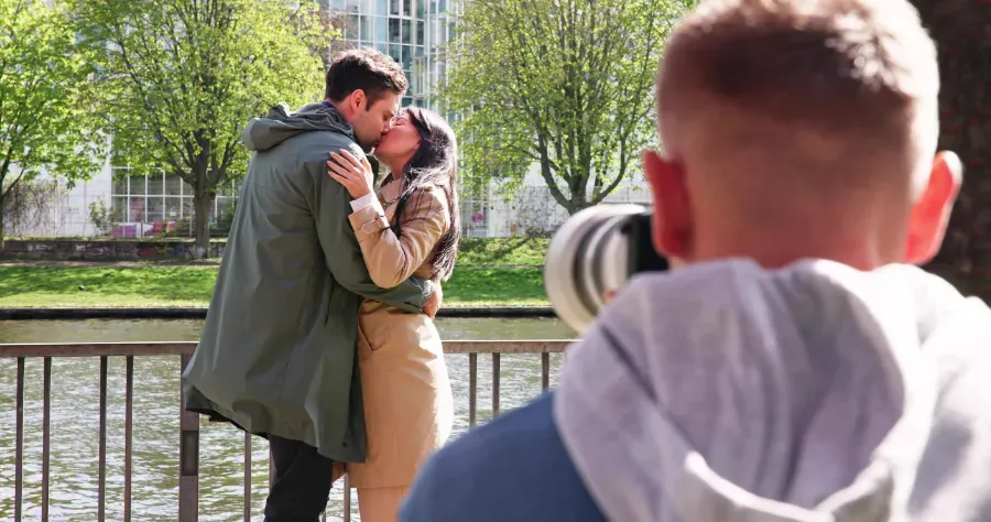 Pareja besándose junto a una barandilla a la orilla del río, con un fotógrafo borroso en primer plano.