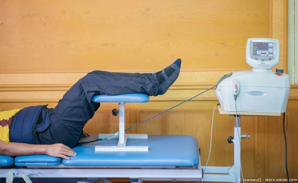 Patient trainiert Bein auf einer Therapieliege neben einem medizinischen Gerät in einem Behandlungsraum