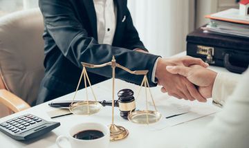 Un abogado le estrecha la mano a un cliente en una mesa con una balanza de justicia y un mazo.