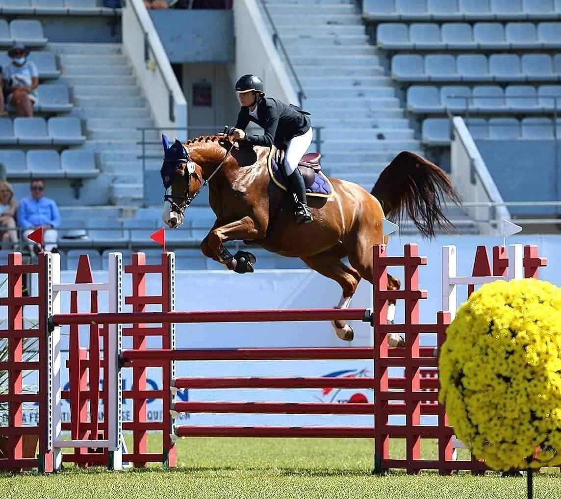Un cheval et son cavalier franchissent un obstacle rouge dans un manège équestre extérieur, avec des gradins bleus en arrière-plan.