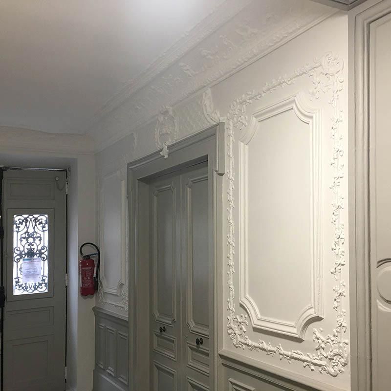 Couloir blanc orné de moulures et de portes grises fermées. Extincteur fixé au mur.