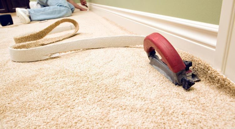 Pose de moquette par une personne utilisant un outil de pose à genou. Moquette beige clair contre une plinthe et un mur blancs.