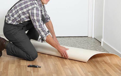 Une personne à genoux pose un revêtement de sol en vinyle. Elle tient le rouleau de vinyle au-dessus du parquet existant. Une porte est visible en arrière-plan.