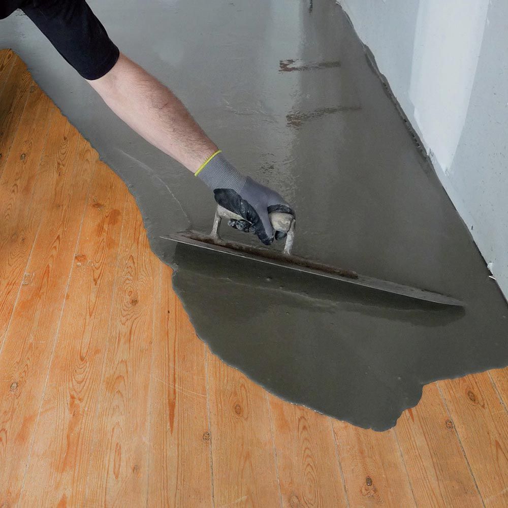 Une personne utilise une truelle pour lisser un enduit de revêtement de sol gris sur un parquet à côté d'un mur blanc.