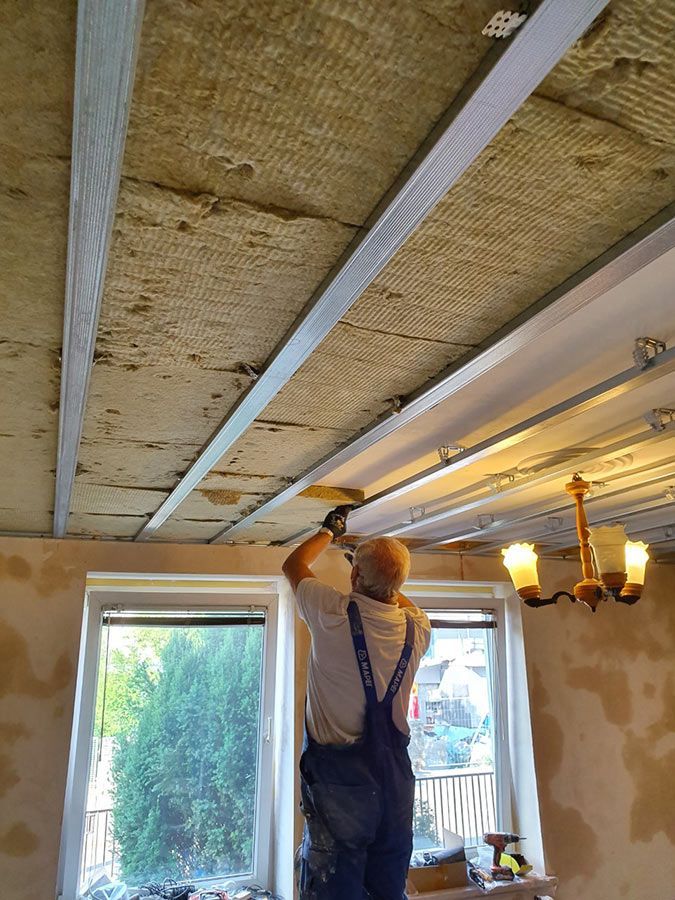 Un homme installe de l'isolant au plafond ; vue intérieure. On aperçoit la charpente, l'isolant et les plaques de plâtre partiellement finies.