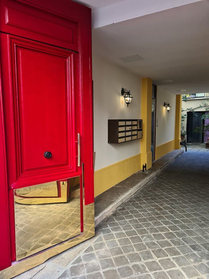Une porte rouge à encadrement doré s'ouvre sur un hall jaune et blanc avec des boîtes aux lettres et un chemin pavé.