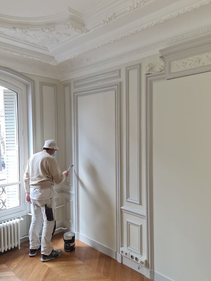 Un peintre en salopette blanche applique de la peinture sur un mur aux boiseries grises dans une pièce avec une fenêtre et un parquet.