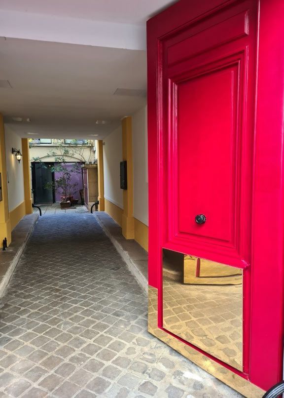 Porte rouge à panneau miroir donnant sur une ruelle pavée aux murs jaunes.