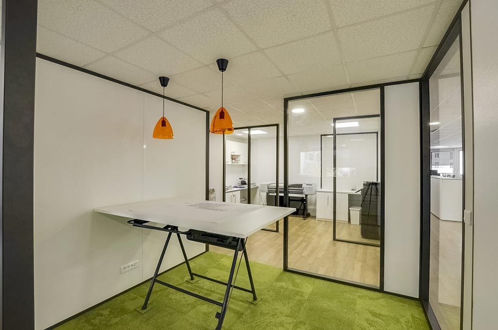 Espace de bureau moderne avec table à dessin, moquette verte et pièces aux parois vitrées.