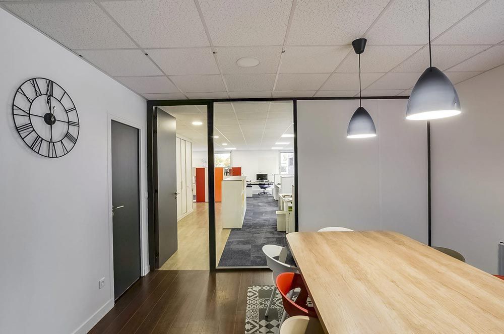 Intérieur de bureau avec horloge, paroi vitrée, table en bois et suspensions lumineuses.
