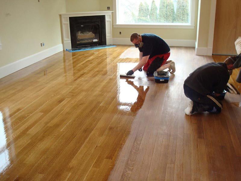 Deux personnes appliquent un produit d'étanchéité sur un parquet brillant dans une pièce avec une cheminée.