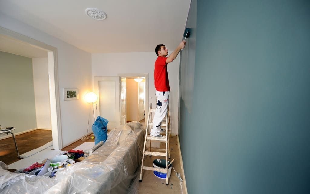 Une personne peint un mur bleu, debout sur une échelle dans une pièce. On aperçoit du matériel de peinture et des meubles recouverts.