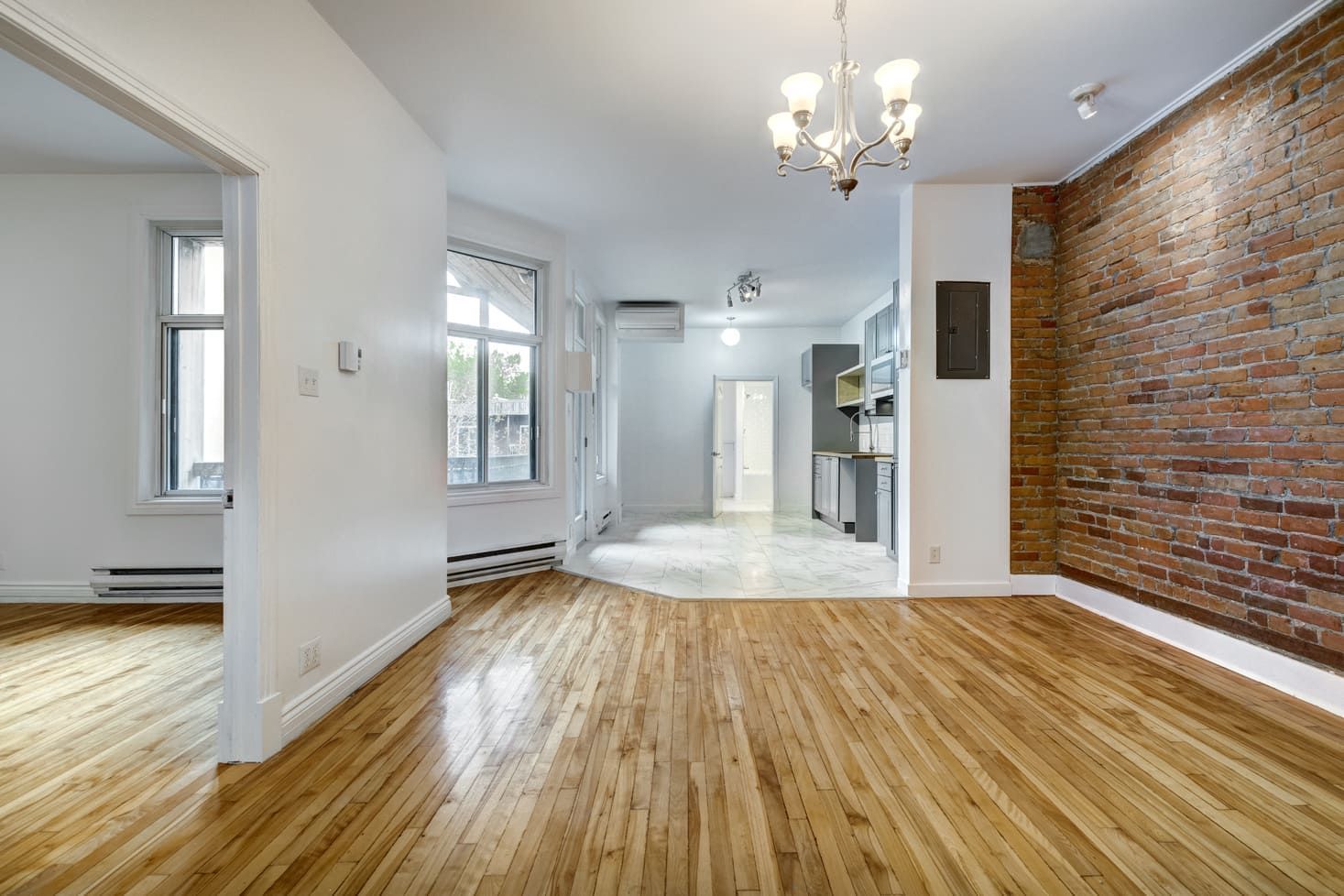 Salon d'appartement vide avec parquet, mur en briques apparentes et lustre.