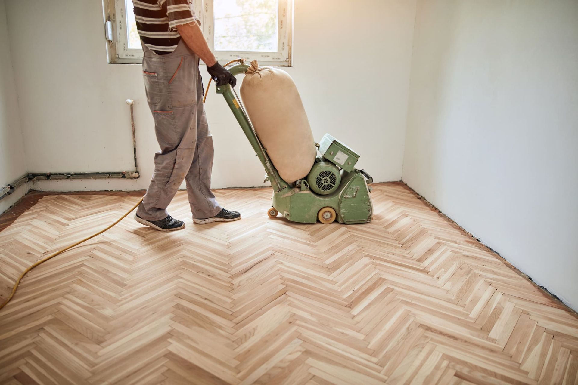 Une personne ponce un parquet à chevrons avec une grande ponceuse industrielle dans une pièce avec une fenêtre.