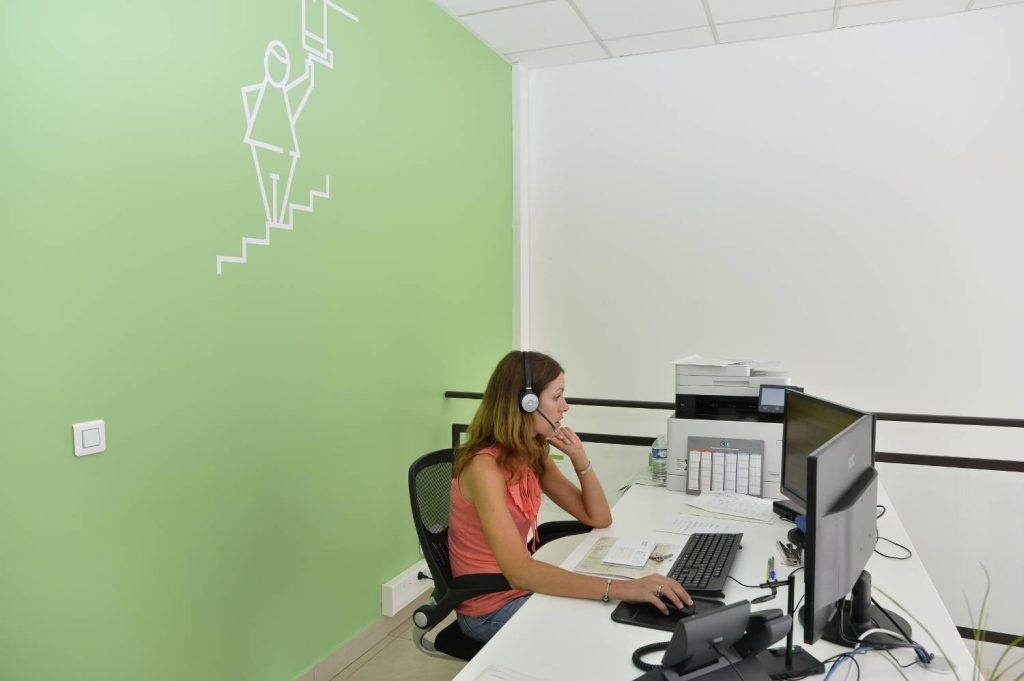 Une femme portant un casque audio travaille à un bureau équipé de deux écrans dans un bureau vert et blanc.