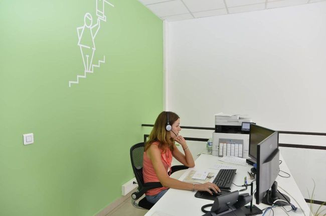 Une femme portant un casque audio travaille à un bureau équipé de deux écrans dans un bureau vert et blanc.