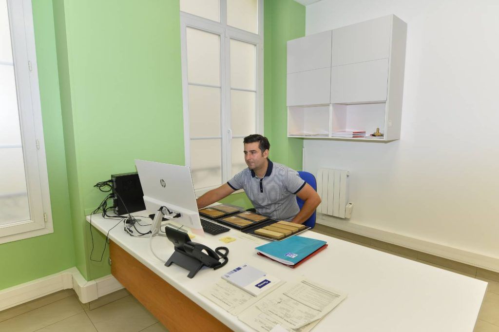 Un homme assis à un bureau avec un ordinateur regarde un écran dans un bureau vert clair et blanc.