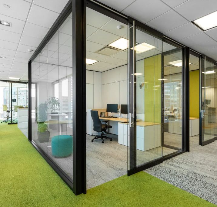 Bureau avec pièces vitrées, moquette verte, bureaux, chaises et lumière naturelle.