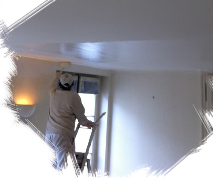 Une personne sur une échelle peint un plafond blanc dans une pièce avec une fenêtre et un luminaire mural.