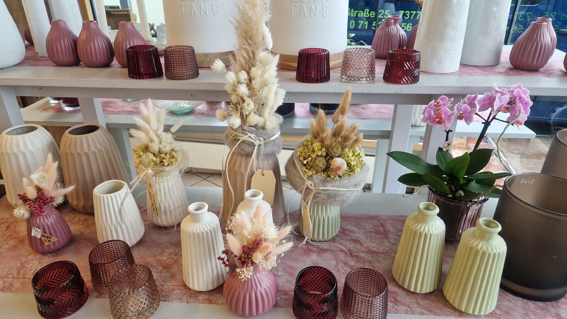 Präsentation von Vasen, Trockenblumen und Kerzenhaltern in Weiß-, Rosa- und Brauntönen auf Regalen.