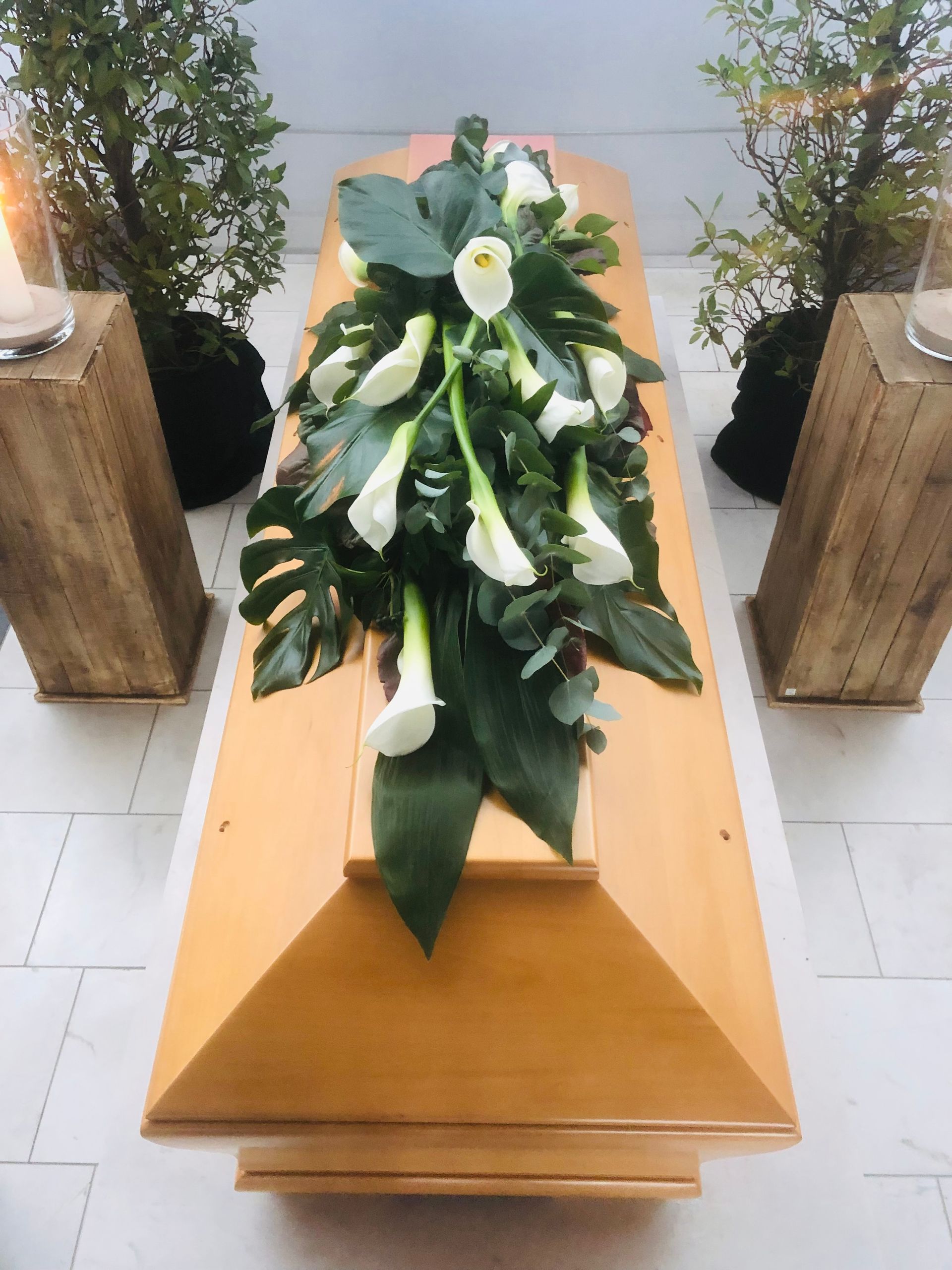 Ein Holzsarg mit weißen Calla-Lilien, Monstera-Blättern und Grünpflanzen in einem hellen Raum.