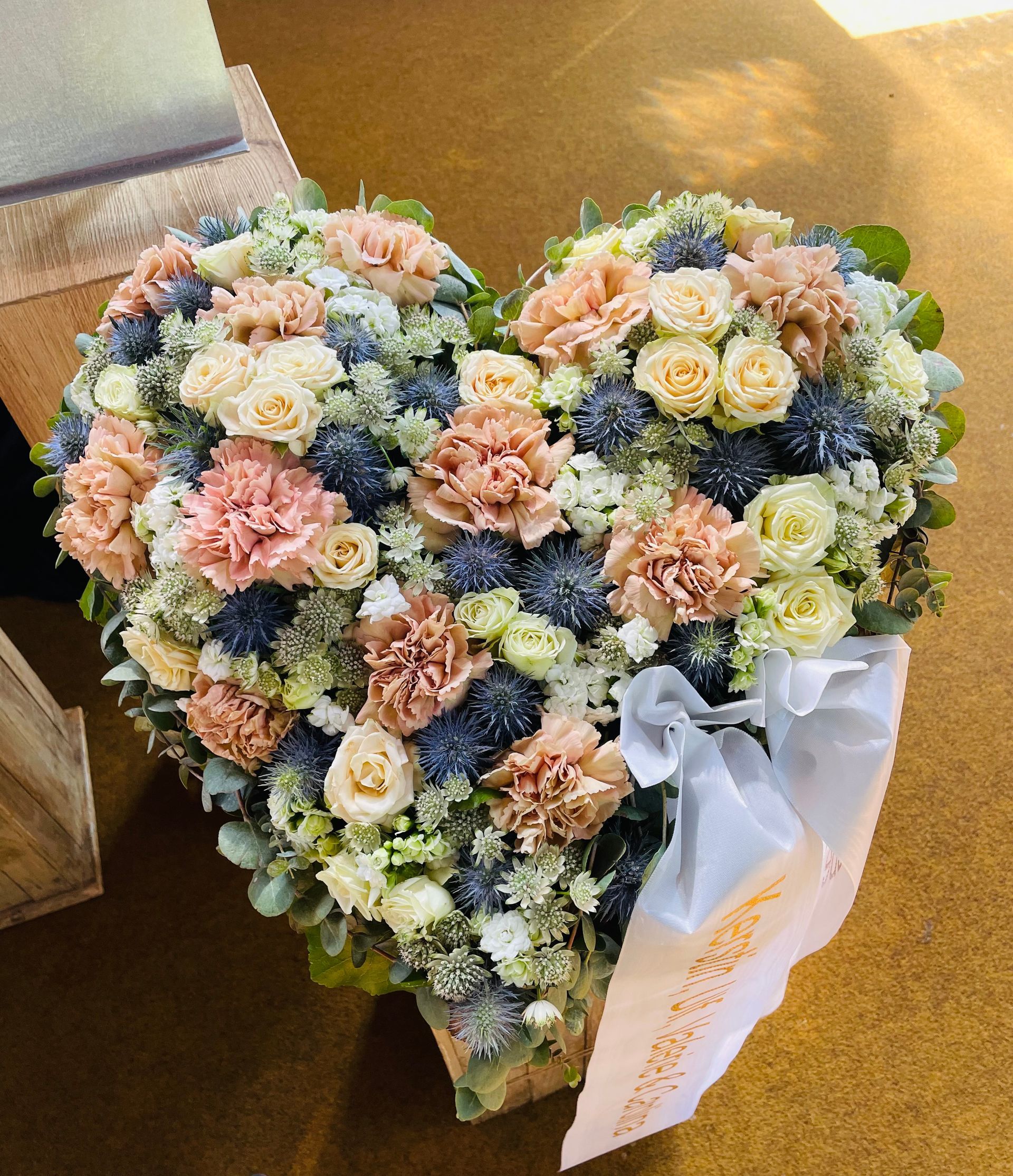 Herzförmiges Blumenarrangement mit pfirsichfarbenen, cremefarbenen und blauen Blumen und einem weißen Band auf einer goldenen Oberfläche.