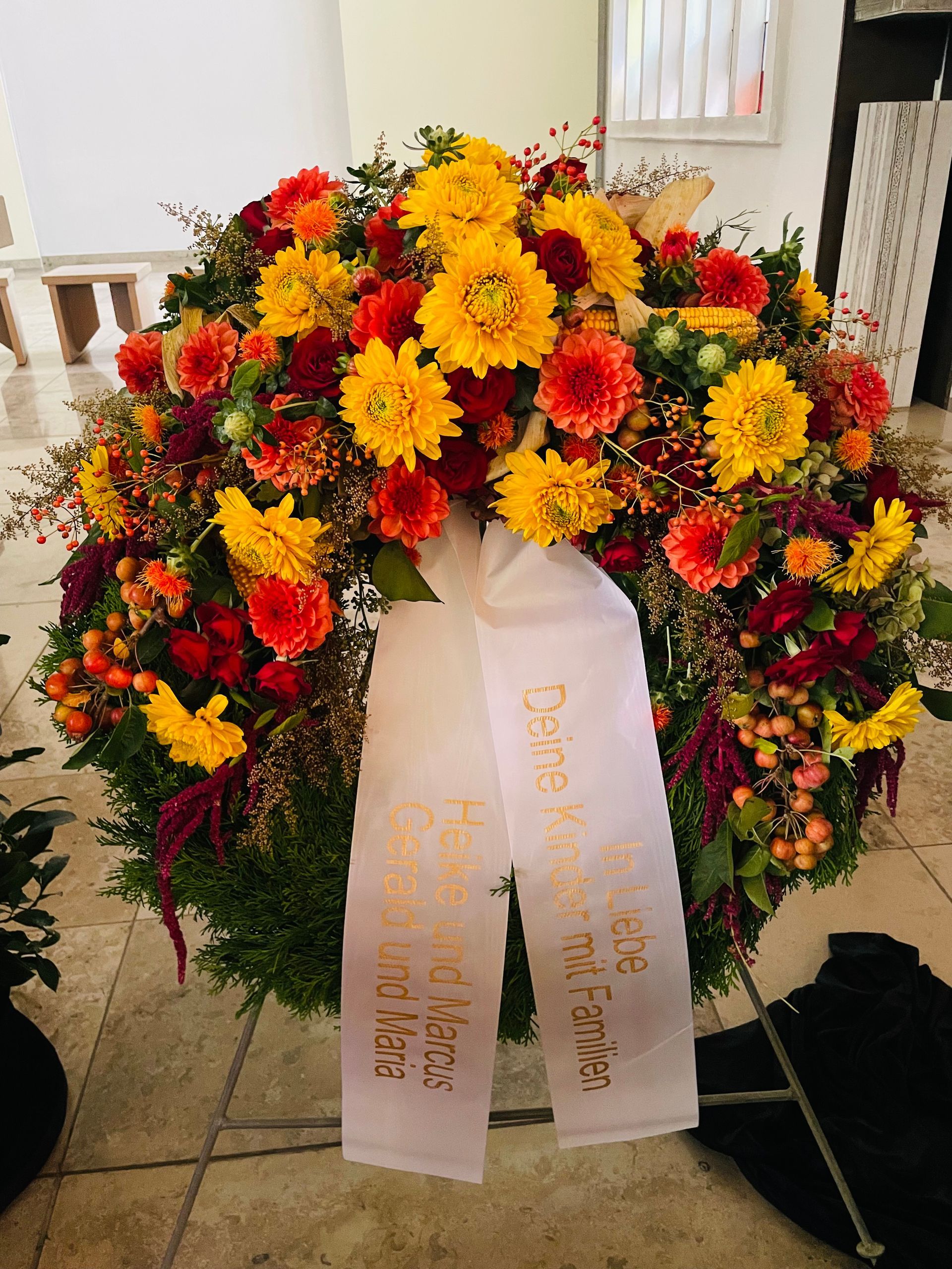 Trauerkranz mit gelben, orangefarbenen und roten Blumen, weißes Band mit goldenem Text.