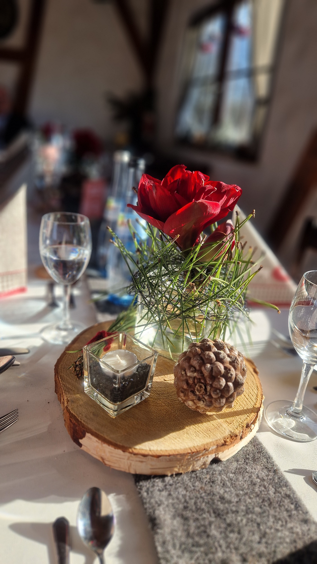 Tafelaufsatz mit roter Rose, Kerze und Tannenzapfen auf einer Holzscheibe, auf einem Tisch mit Gläsern und Besteck gedeckt.
