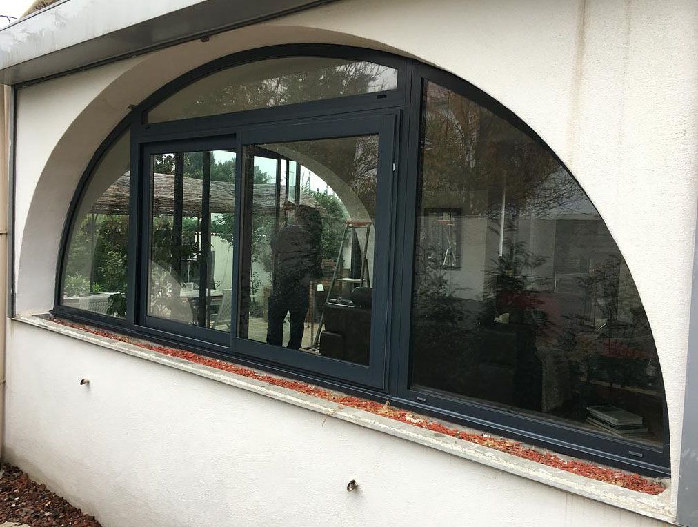 Fenêtre cintrée avec portes coulissantes en verre, cadres foncés, encastrée dans un mur extérieur blanc. Une personne est visible à l'intérieur.
