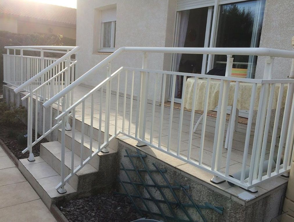 Rambarde en métal blanc sur les marches menant à une porte-fenêtre donnant sur le patio. Une table blanche avec une serviette est posée sur le patio.