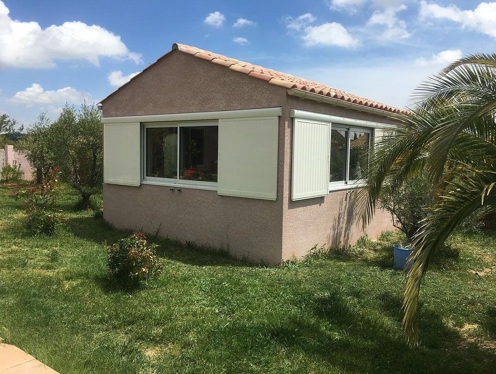 Petit bâtiment en stuc avec des volets blancs aux fenêtres, sur une pelouse herbeuse sous un ciel partiellement nuageux.