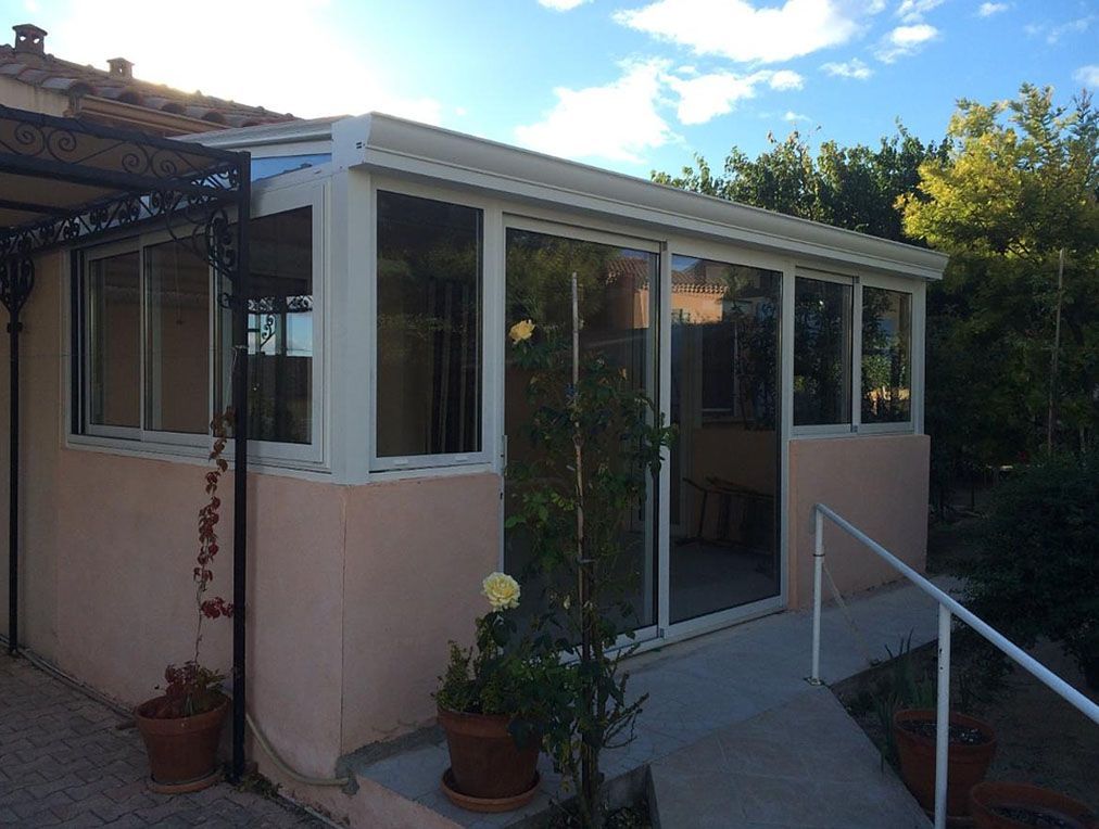 Extension de type véranda avec fenêtres en verre et encadrement blanc, attenante à un bâtiment en stuc rose.