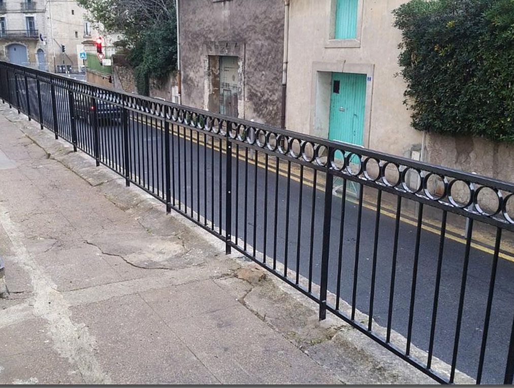 Rambarde en métal noir le long d'un trottoir, donnant sur une rue bordée d'immeubles et d'une porte turquoise.
