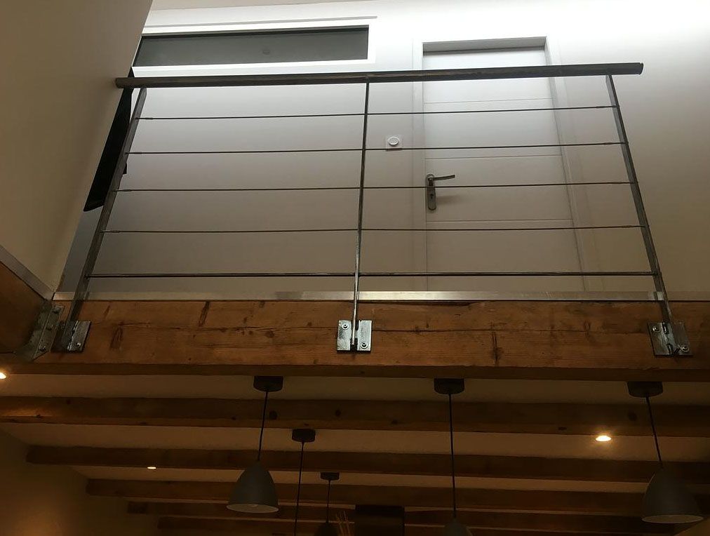 Rambarde de mezzanine composée de barres d'acier horizontales, fixée à une poutre en bois. Porte et petite fenêtre visibles en arrière-plan.