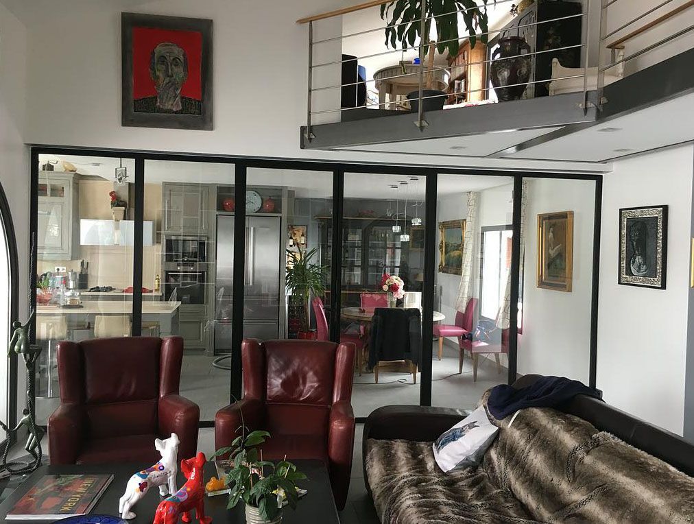 Salon avec paroi vitrée donnant sur la cuisine ; fauteuils rouges, canapé, œuvres d'art et mezzanine.