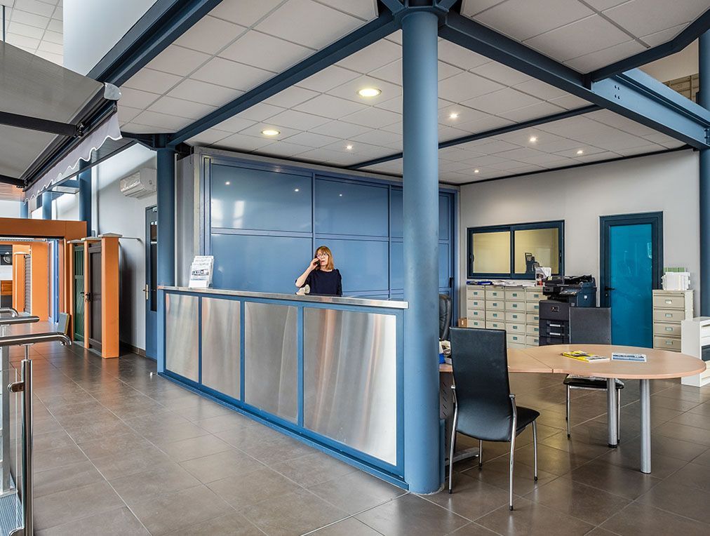 Espace d'accueil avec une femme au téléphone derrière un comptoir argenté, un mur bleu et des poutres de soutien.