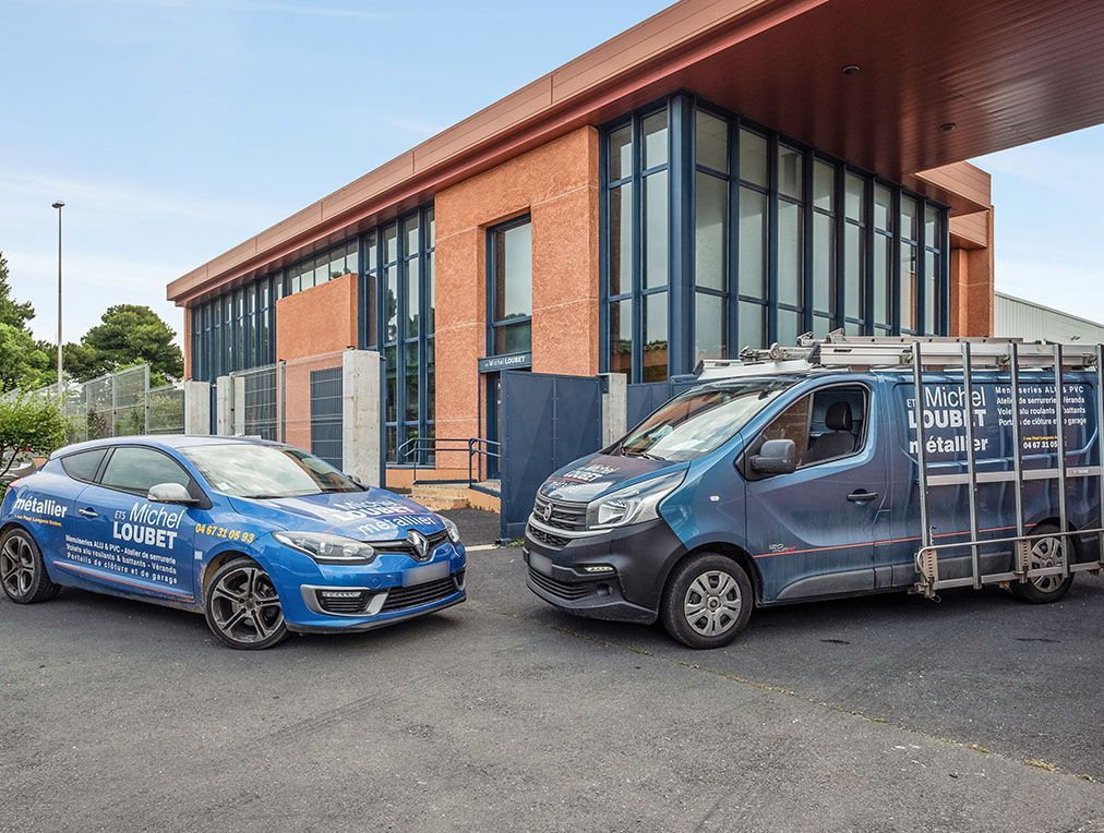Une voiture et une camionnette bleues sont garées devant un bâtiment en briques. La camionnette est équipée d'un porte-verres sur le toit et les voitures arborent des logos d'entreprises.