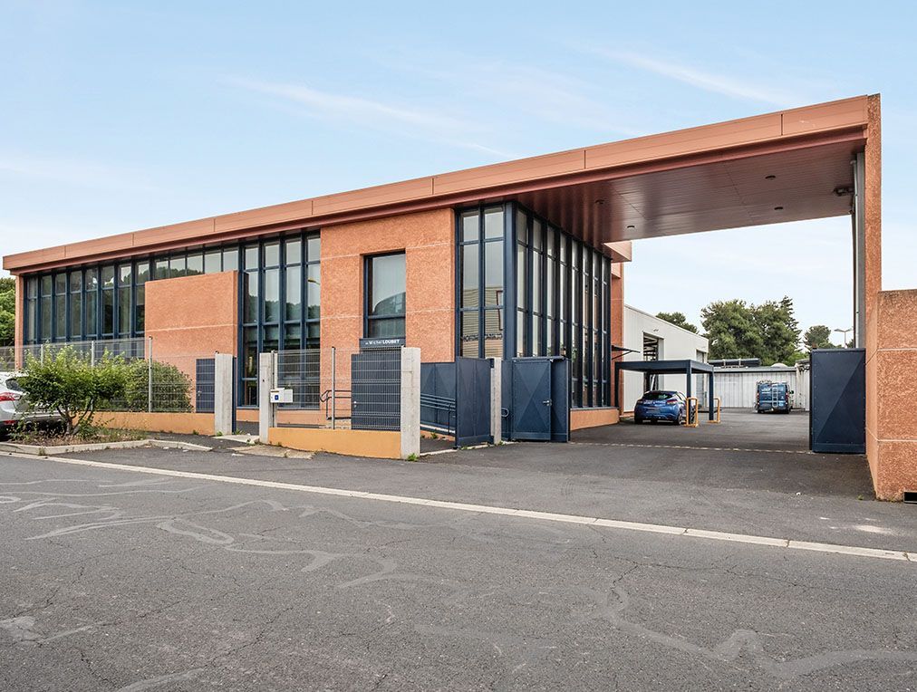 Bâtiment en briques avec de grandes fenêtres et une entrée voûtée. Des voitures garées sont visibles.