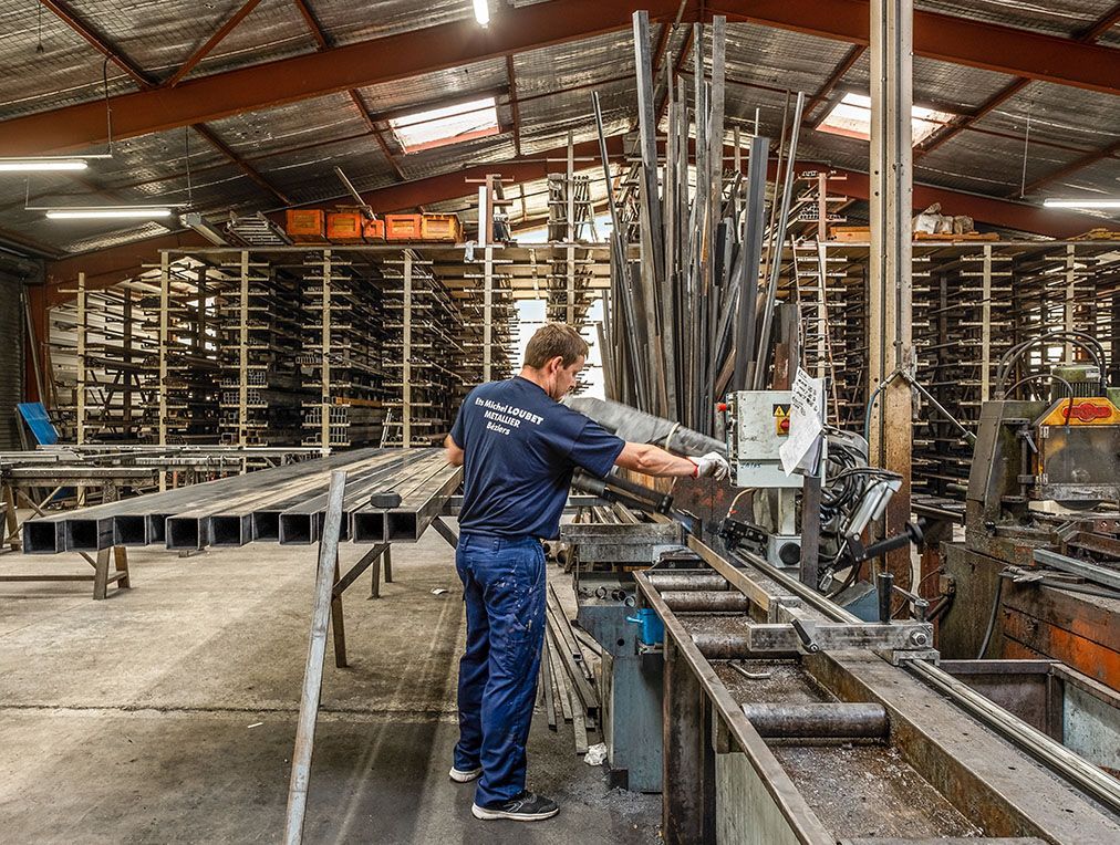 Un ouvrier utilise une scie pour couper des tubes métalliques dans un atelier ; des piles de métal sont visibles en arrière-plan.