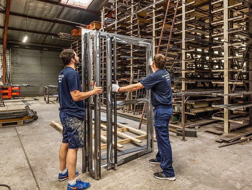 Deux ouvriers manipulent une structure métallique dans un entrepôt. Des étagères remplies d'articles métalliques sont visibles en arrière-plan.