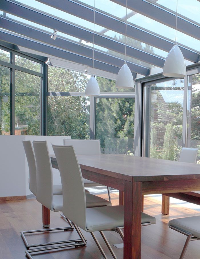Salle à manger avec table en bois, chaises blanches, fenêtres en verre et suspensions lumineuses.