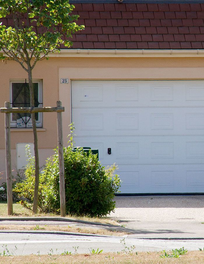 Garage avec une porte blanche à côté d'un petit arbre et de buissons verts devant un bâtiment couleur pêche clair.