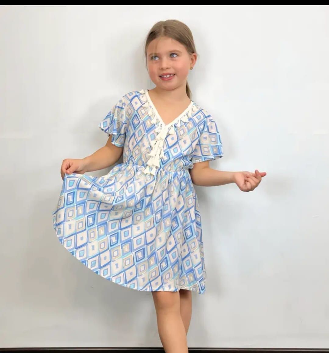 Una niña con un vestido azul y blanco está parada frente a una pared blanca.