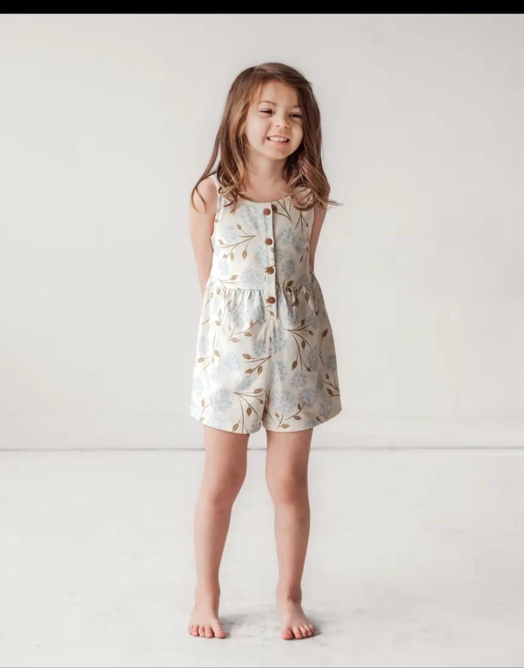 Una niña que lleva un vestido blanco y pantalones cortos está parada frente a una pared blanca.