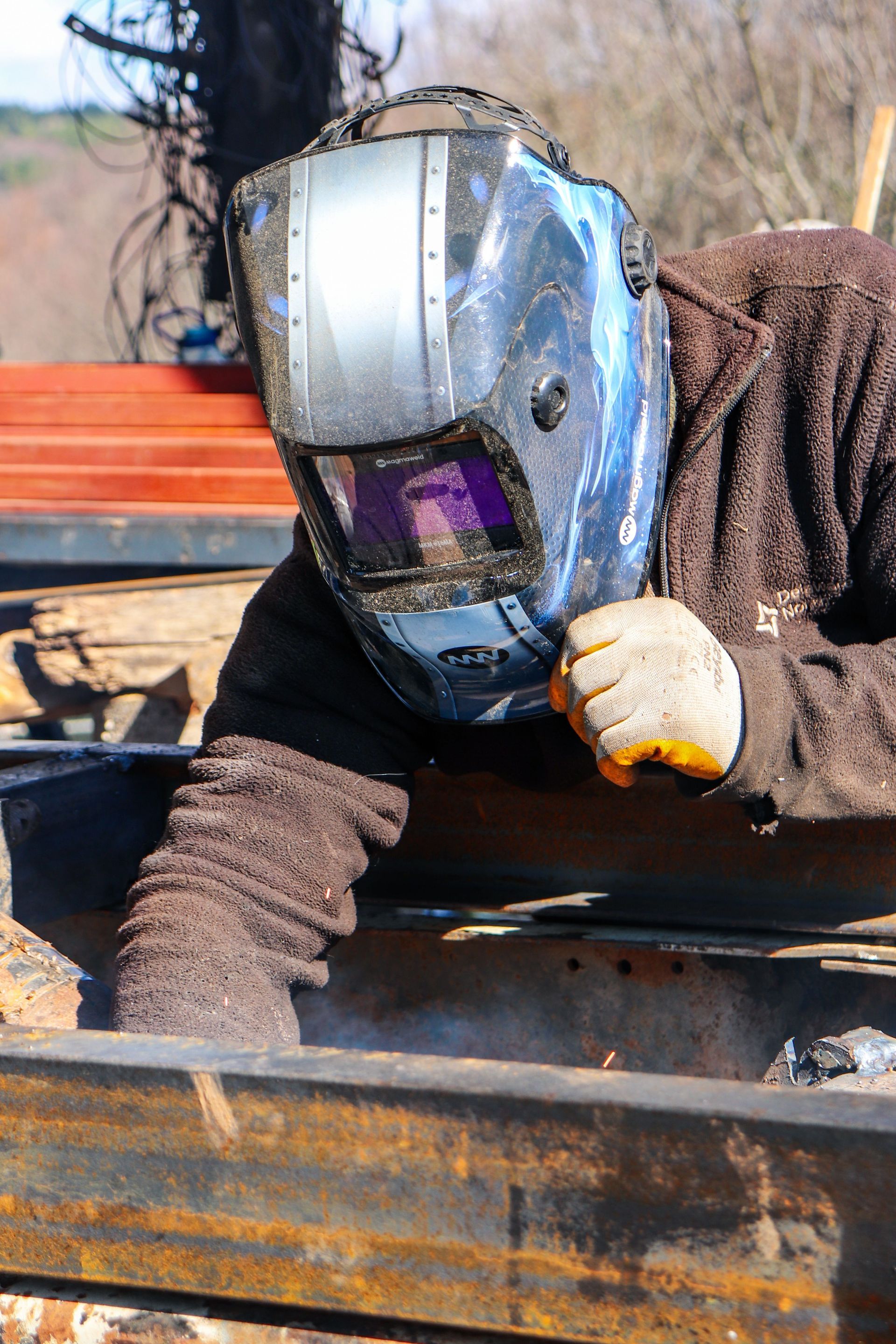 Un soudeur portant un casque de protection se penche sur une poutre métallique à l'extérieur.