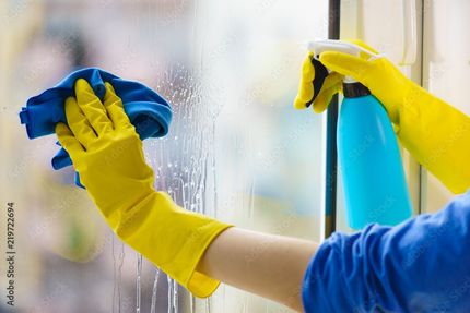 Des mains gantées de jaune nettoient une vitre avec un chiffon bleu et un vaporisateur.